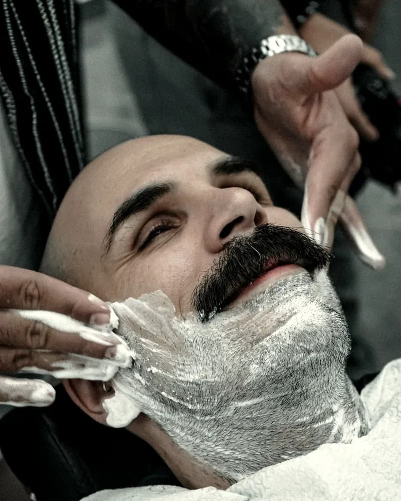 Cliente reclinado con espuma de afeitar mientras el barbero perfila la barba